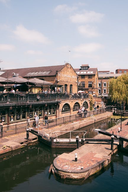 A large, weathered metal railway bridge painted in shades of teal and rust, spanning across an urban street in Camden. The bridge features prominent yellow lettering spelling 'Camden Lock' with a smal